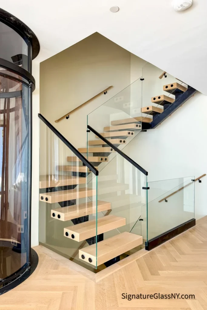 Clear glass stair railings with black handrails beside floating wood treads and a curved glass enclosure.